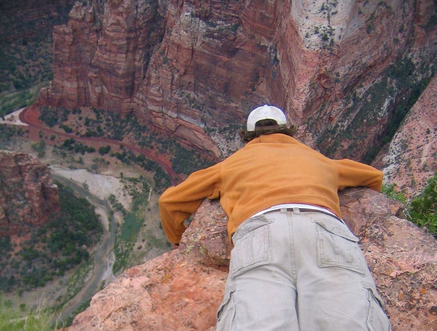 A person leans over a rocky ledge to look down into a deep canyon.