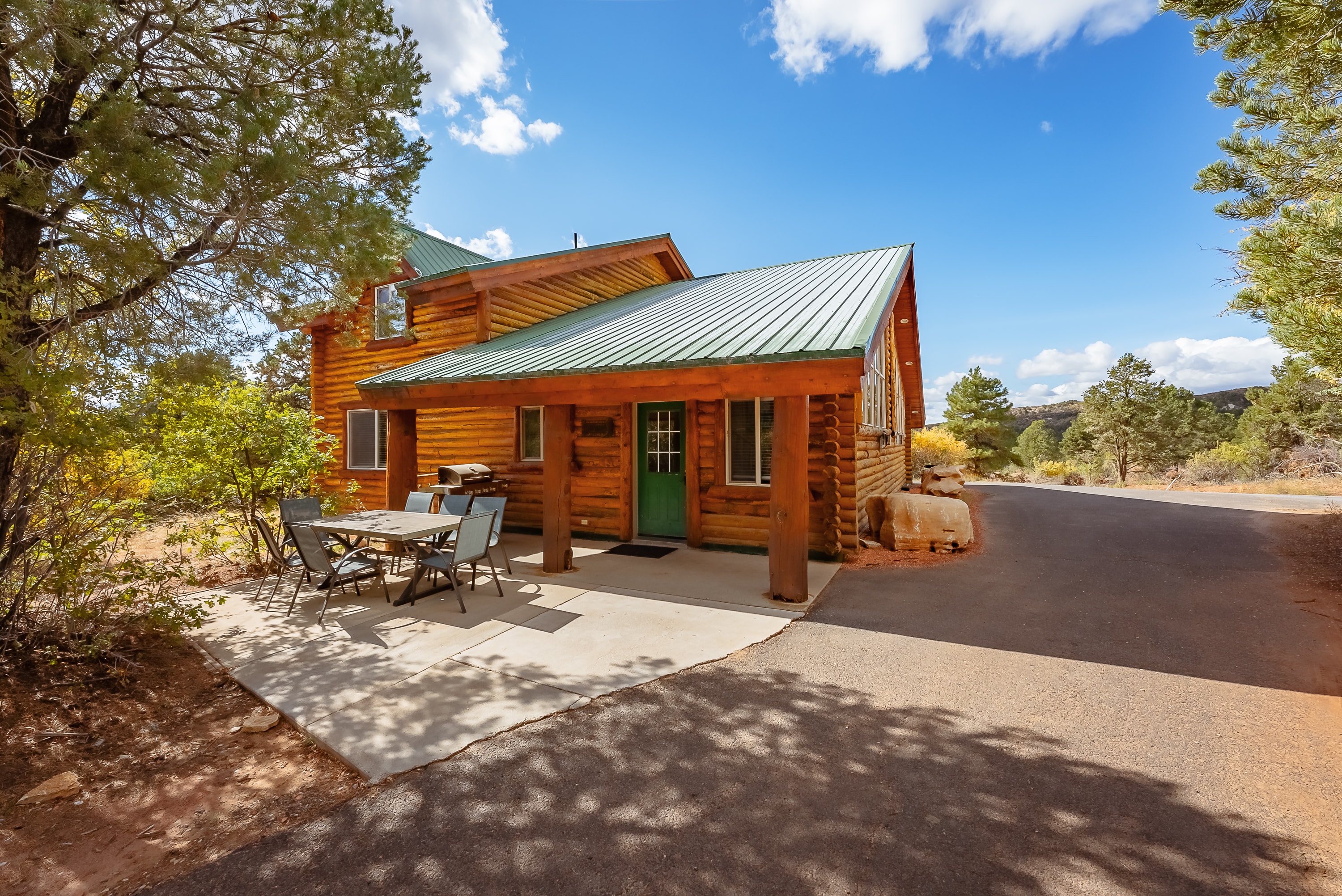 A log cabin with a green metal roof, surrounded by trees, featuring a patio and outdoor seating area.