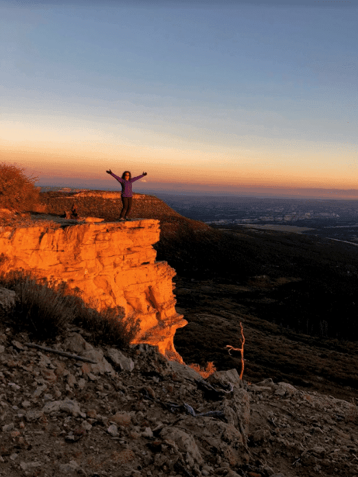 A person stands with arms outstretched on a rocky ledge during sunset, overlooking a vast landscape.