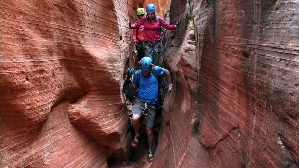 Three climbers navigating a narrow slot canyon with reddish rock formations.