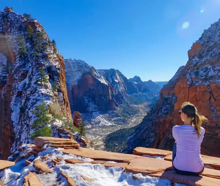 A person sits on a snowy cliff, overlooking a vast canyon surrounded by mountains under a clear blue sky.