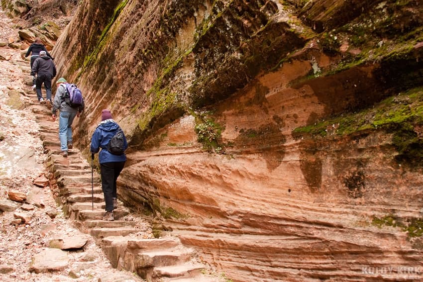 Hikers ascend a rocky path carved into a steep, layered canyon wall.
