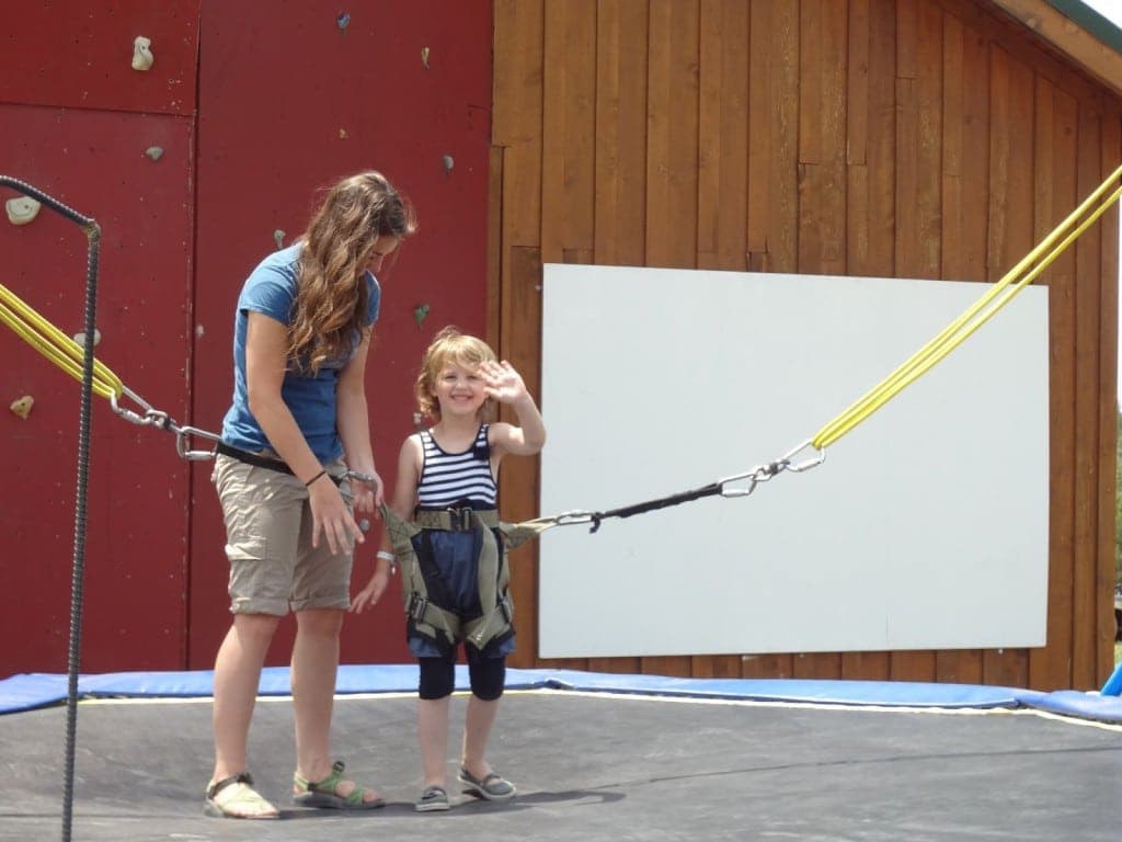 A smiling child in a harness waves at the camera while standing next to an adult on a trampoline.