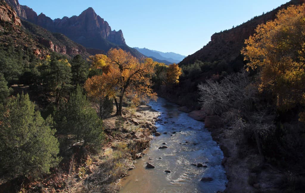 A serene river flows through autumn-colored trees with rugged mountains in the background.
