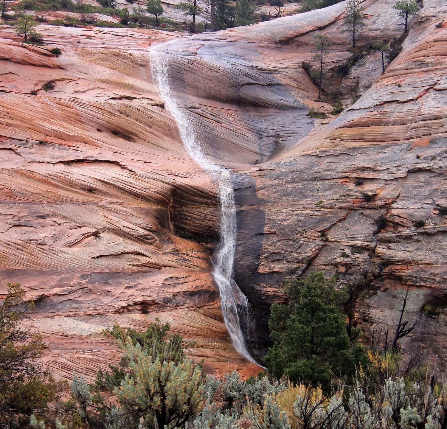 A small waterfall cascading down smooth, multicolored rock formations surrounded by sparse greenery.