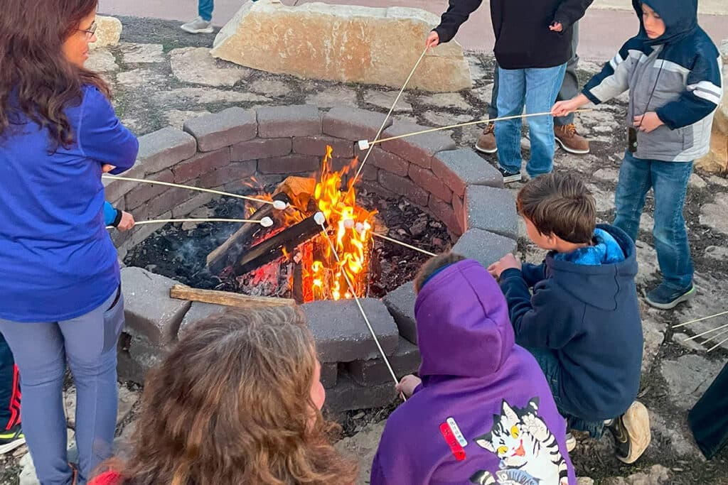 A group of children roast marshmallows over a campfire in a stone fire pit.