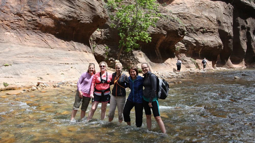 A group of five women stands in a shallow river, smiling, with rocky cliffs in the background.
