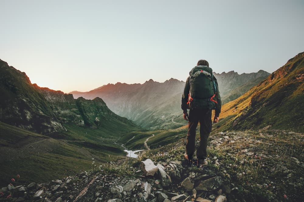 A hiker stands on a mountain trail, overlooking a valley at sunset.