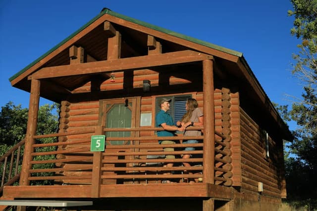 A couple stands on the porch of a log cabin under a clear blue sky.