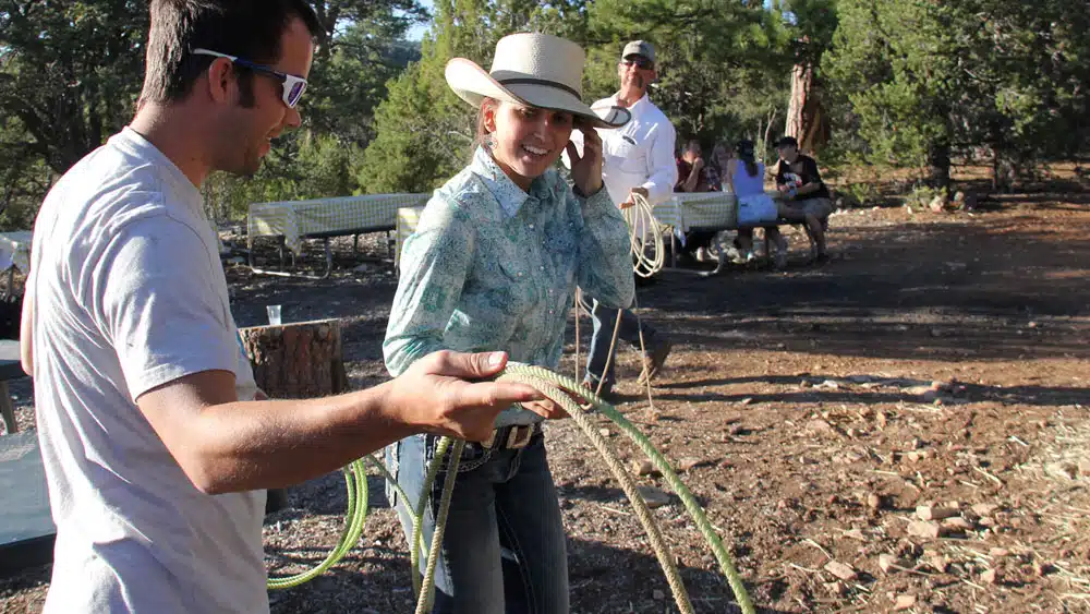 A woman in a cowboy hat smiles as she adjusts her hair while a man demonstrates rope techniques in a rustic outdoor setting.