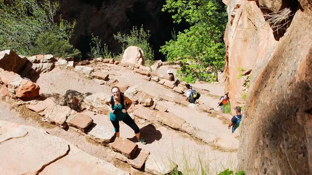 A hiker pauses on a rocky, ascending trail surrounded by trees and other hikers in the background.