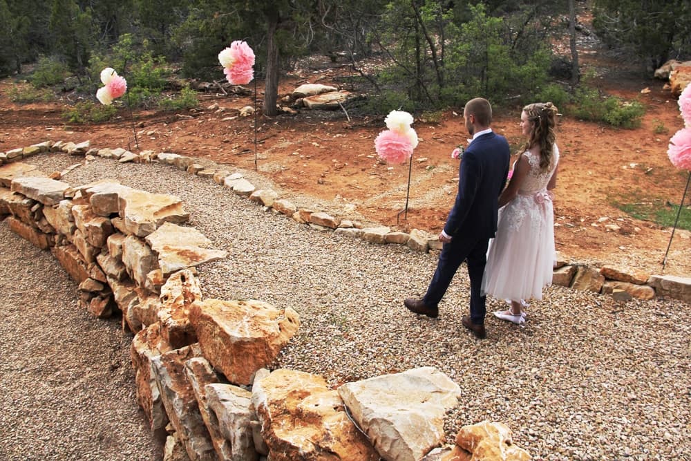 A couple walks along a stone pathway adorned with pink floral decorations in a natural setting.