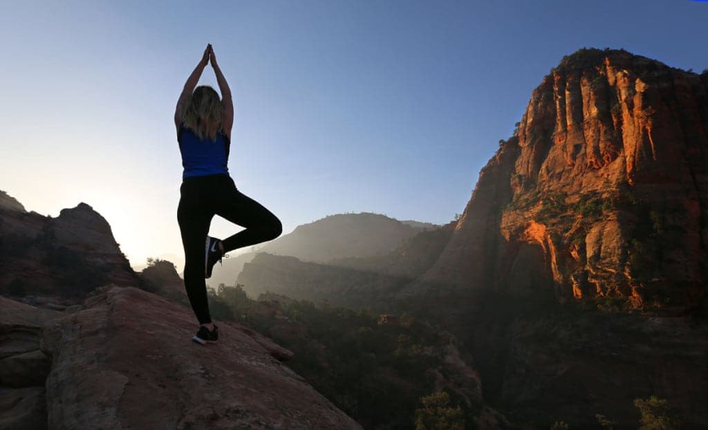 A person practicing yoga on a rock ledge with mountains in the background during sunrise.