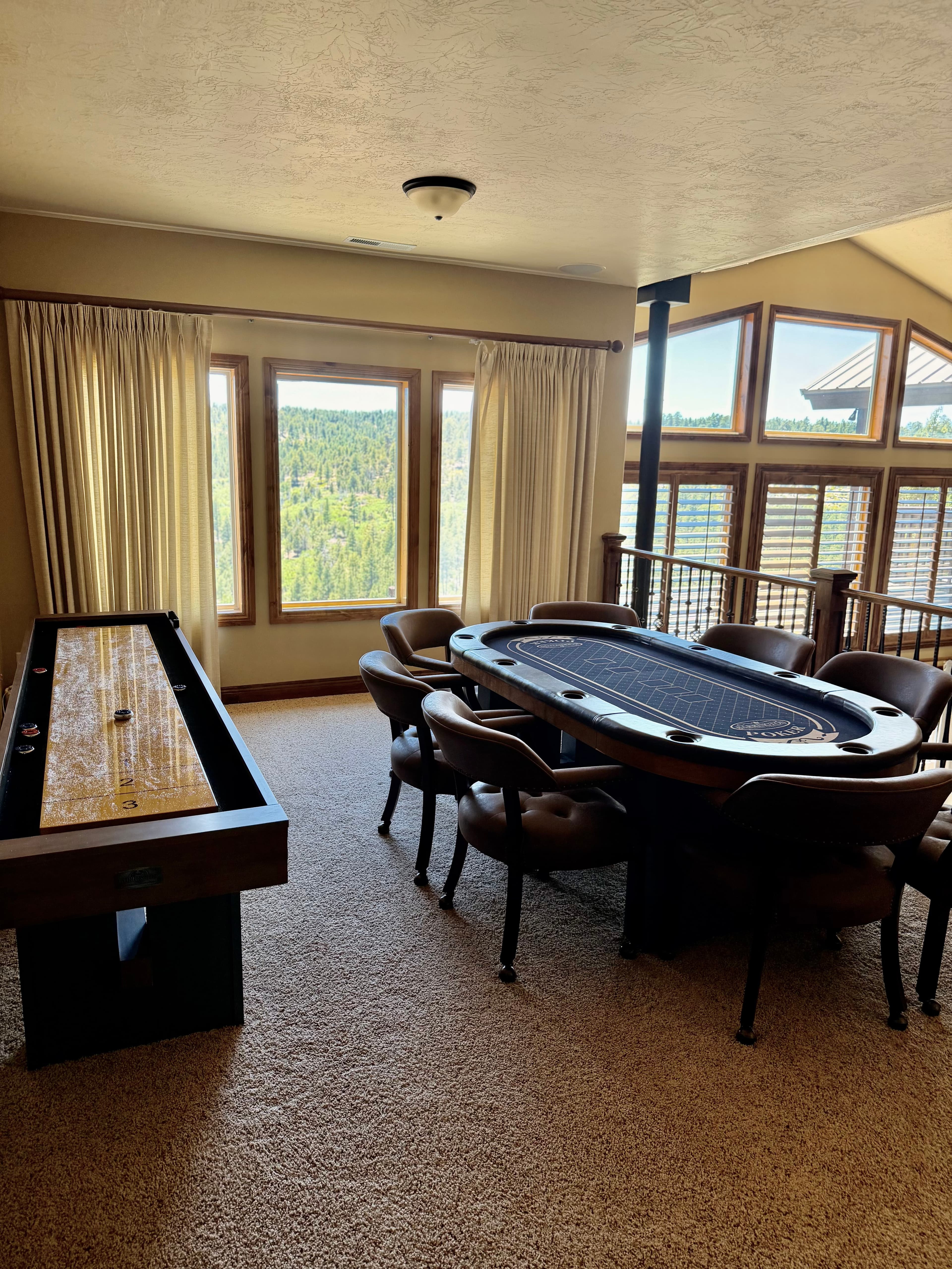 A gaming room features a poker table and a shuffleboard, with large windows showcasing a forest view.