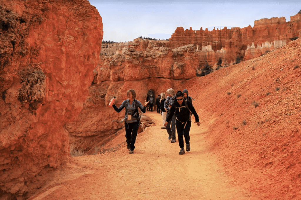 Hikers traverse a narrow, sandy path surrounded by red rock formations in a canyon.