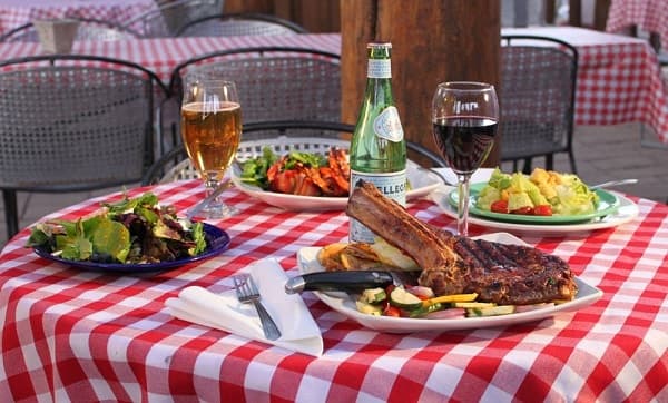 A table set with a variety of dishes including a rib steak, salads, and drinks, all arranged on a red-checked tablecloth.