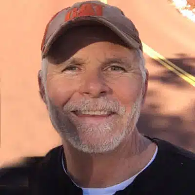 A smiling man with a gray beard and a cap stands outdoors.