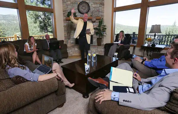 A group of six professionals engage in a meeting in a spacious, well-lit room with large windows and a stone fireplace.
