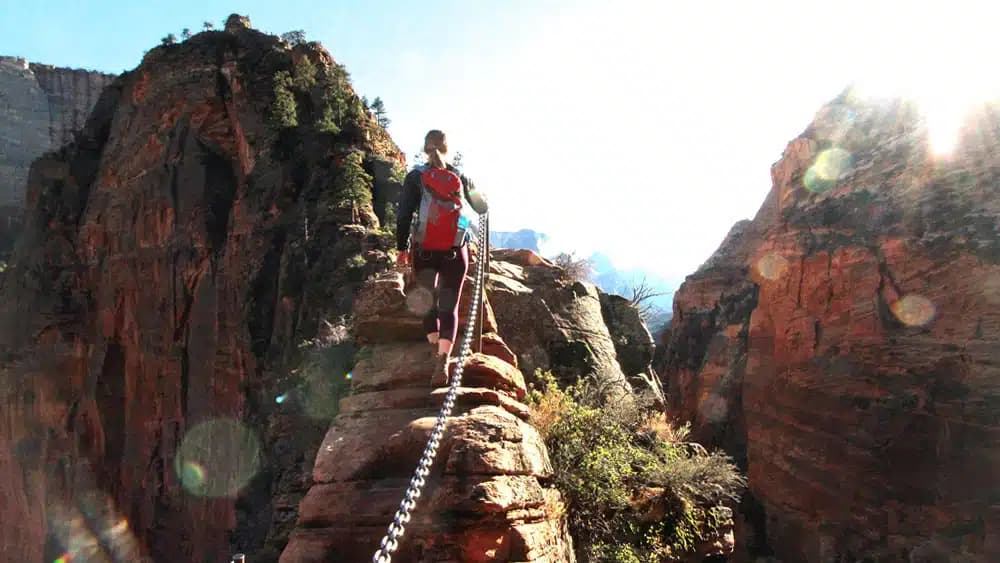 A hiker climbs a narrow rock ledge with a chain for support, surrounded by tall cliffs and bright sunlight.