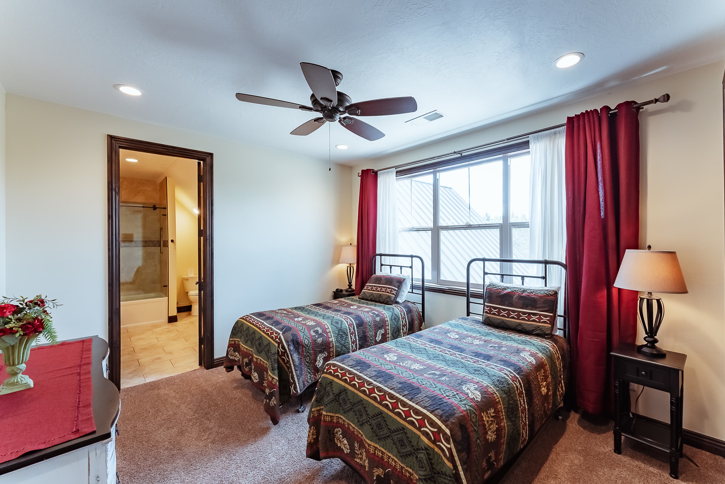 A cozy bedroom with two twin beds, a window with red curtains, and a door leading to an attached bathroom.
