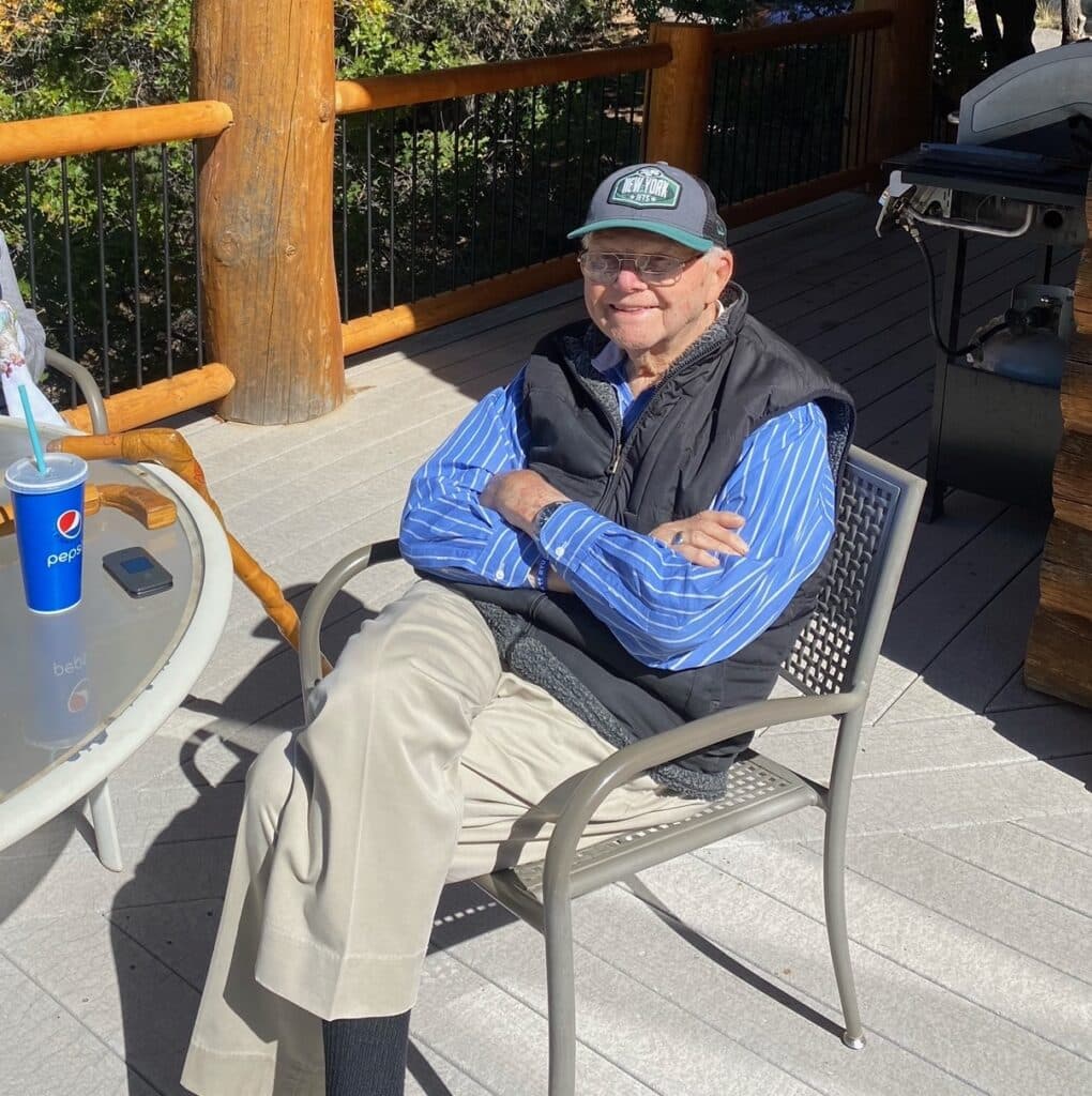 An elderly man sits comfortably on a patio chair, wearing a cap and a blue striped shirt, with a drink on the table beside him.