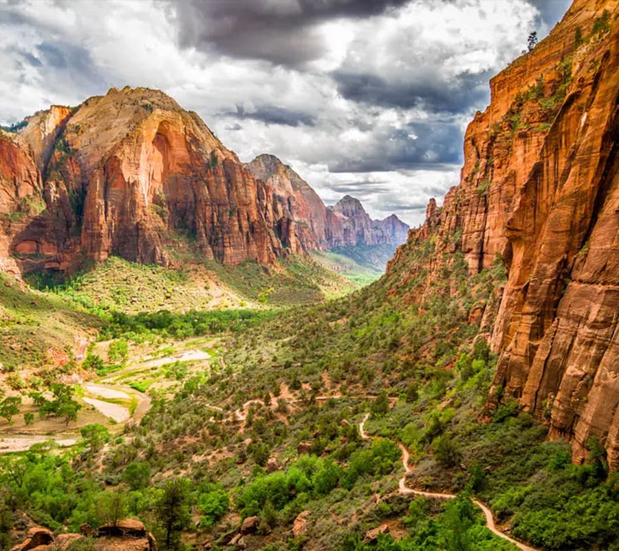 A sweeping view of a canyon surrounded by towering red cliffs and lush greenery under a cloudy sky.