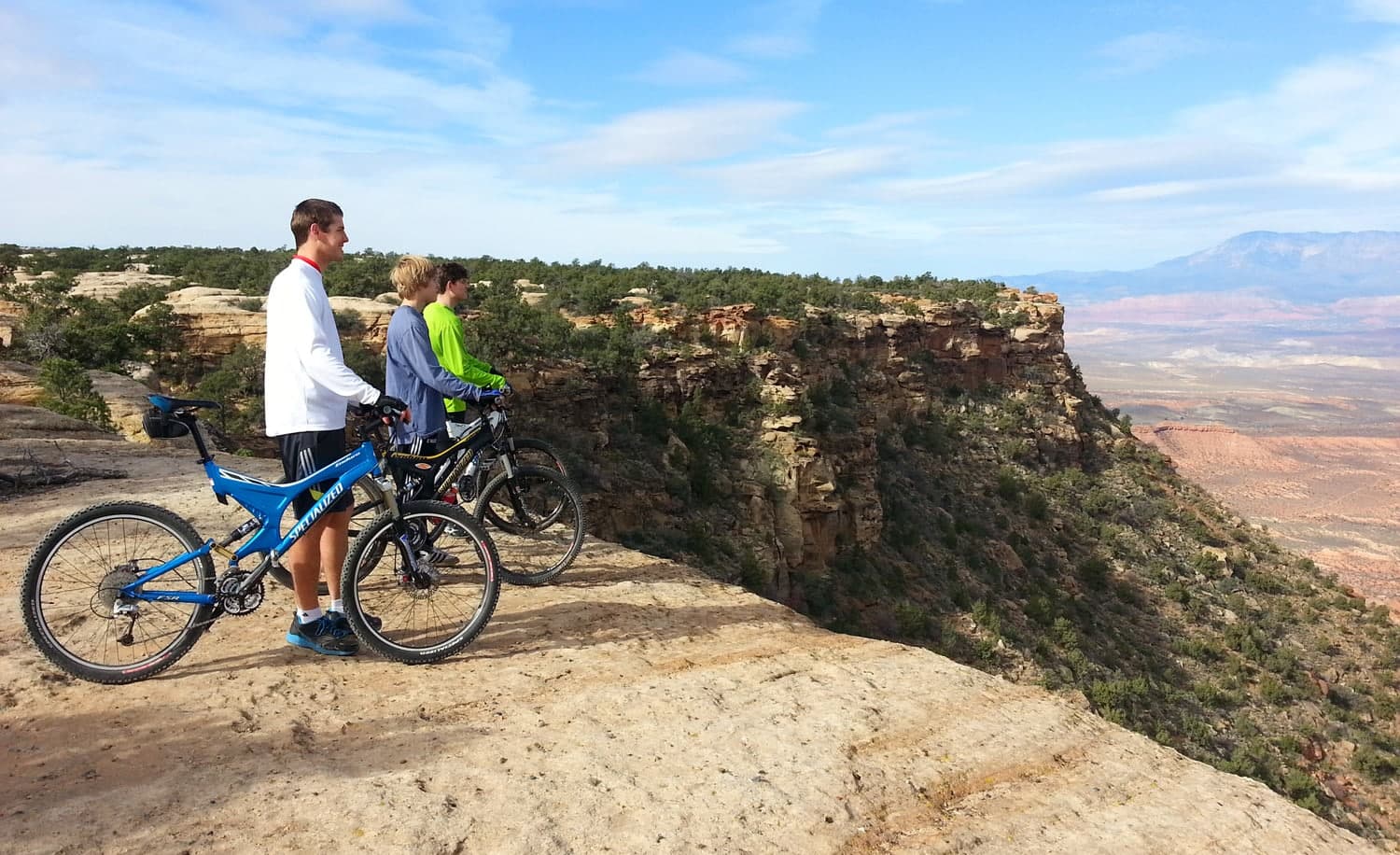 Three cyclists stand at the edge of a rocky cliff overlooking a vast landscape.