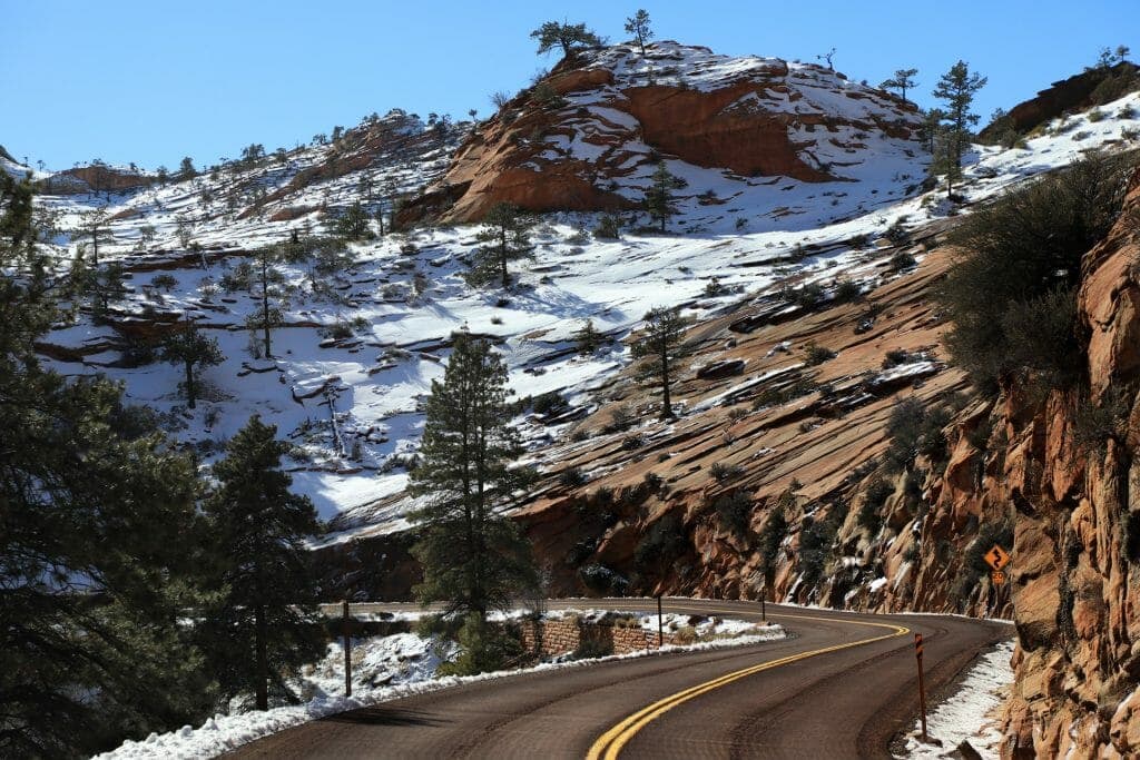 Curving road through a snowy landscape with rocky hills and evergreen trees.