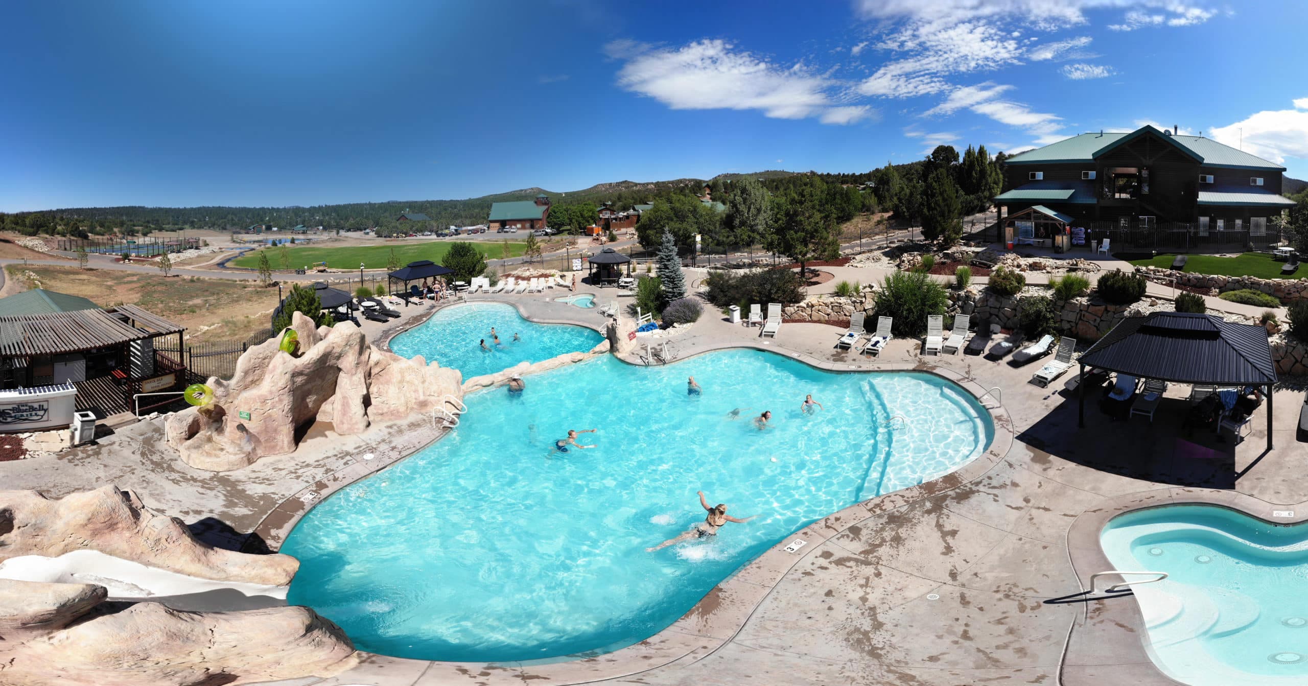 A panoramic view of a resort pool area with people swimming and lounging under a clear blue sky.