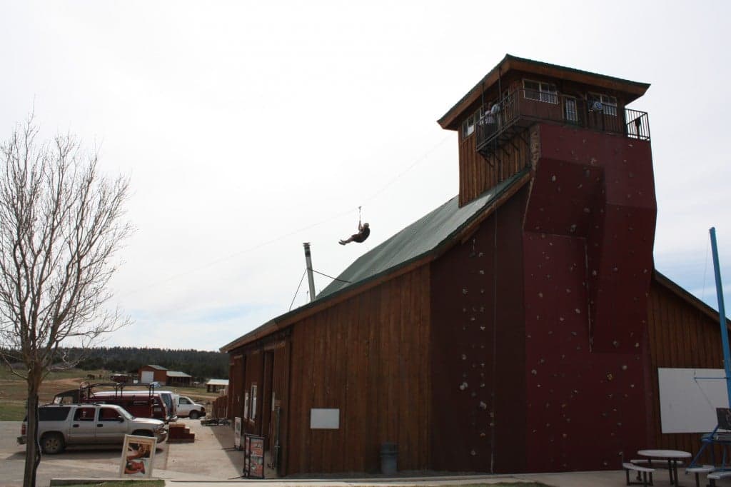 A person zip-lines off a tower next to a wooden building with climbing walls.