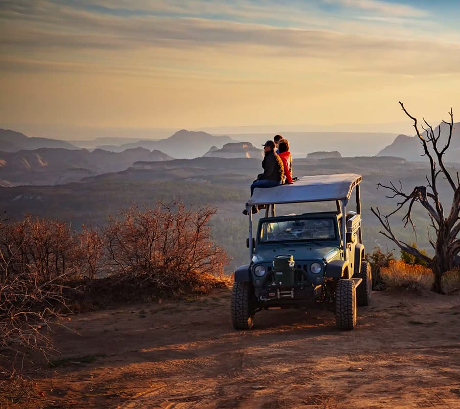 A couple sits on top of a jeep, overlooking a scenic landscape at sunset.