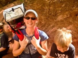 A smiling woman in sunglasses carries a child in a backpack harness while two other children sit nearby.