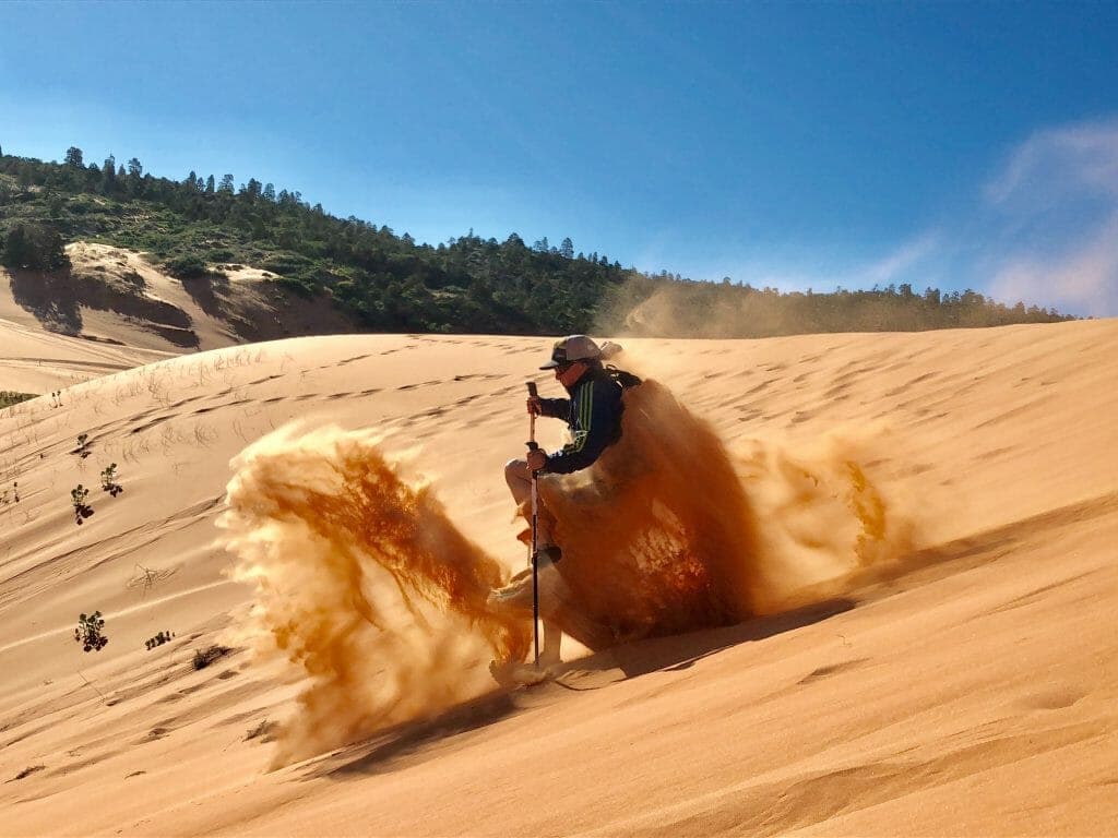 A person skiing down a sandy dune, kicking up a spray of sand against a blue sky.