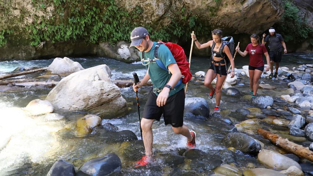 A group of four hikers crosses a shallow river with smooth stones and lush greenery in the background.
