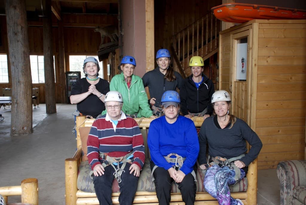 A group of seven people wearing helmets is seated on a rustic wooden couch in a large indoor space.