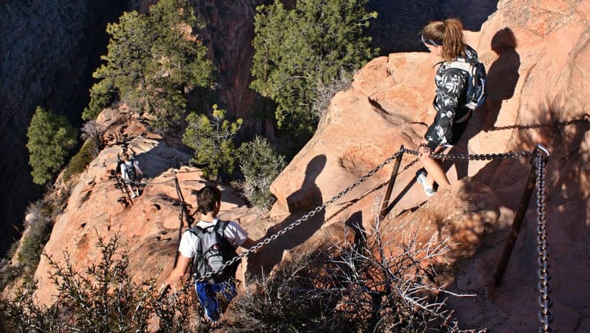 Hikers navigate a rocky trail with a chain railing in a canyon landscape.