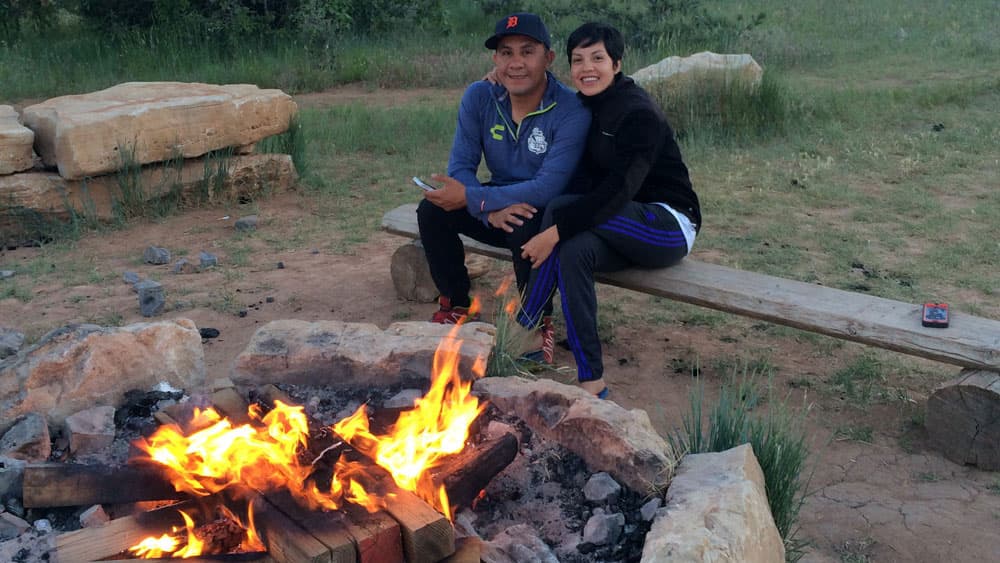 A couple sits on a bench beside a campfire in a grassy outdoor setting.