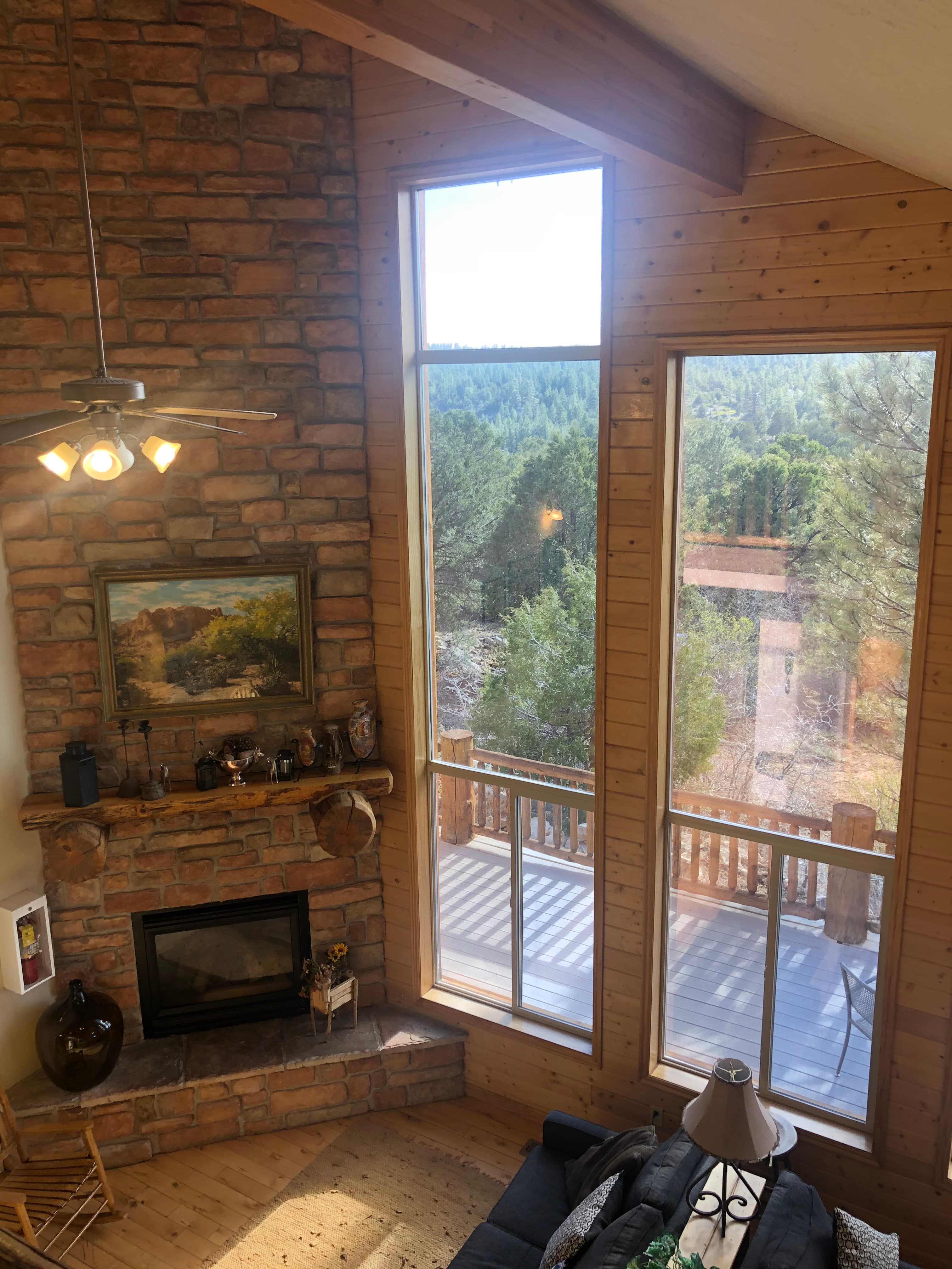 A cozy living room with a stone fireplace, large windows showcasing a forested view, and a balcony.