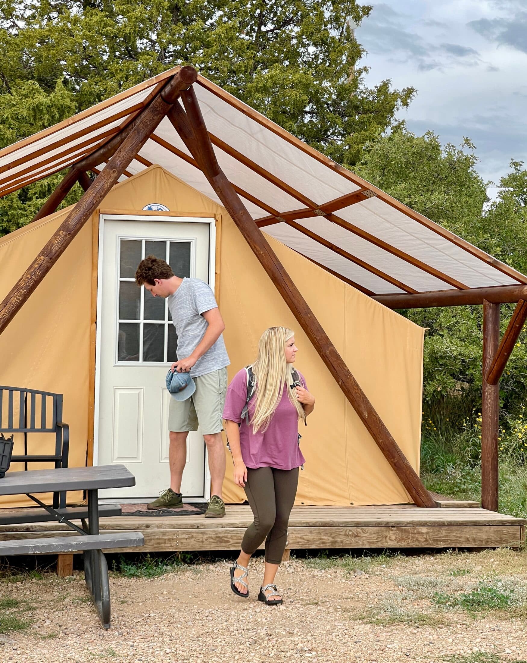 A man and a woman stand outside a tan tent cabin, with the man looking down and the woman wearing a backpack.
