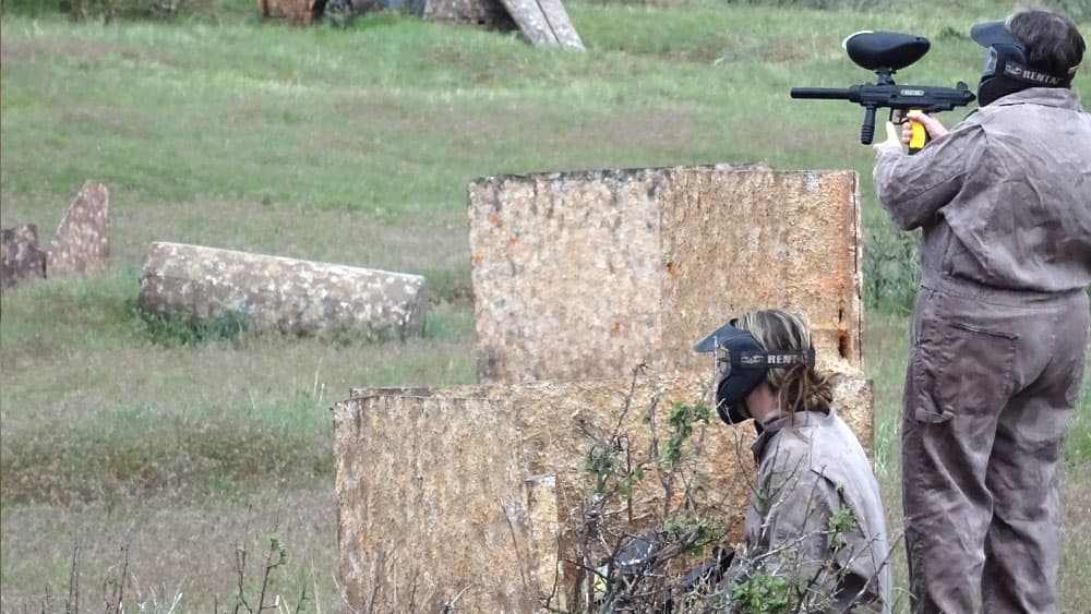 Two players in paintball gear take cover behind barriers in a grassy field.