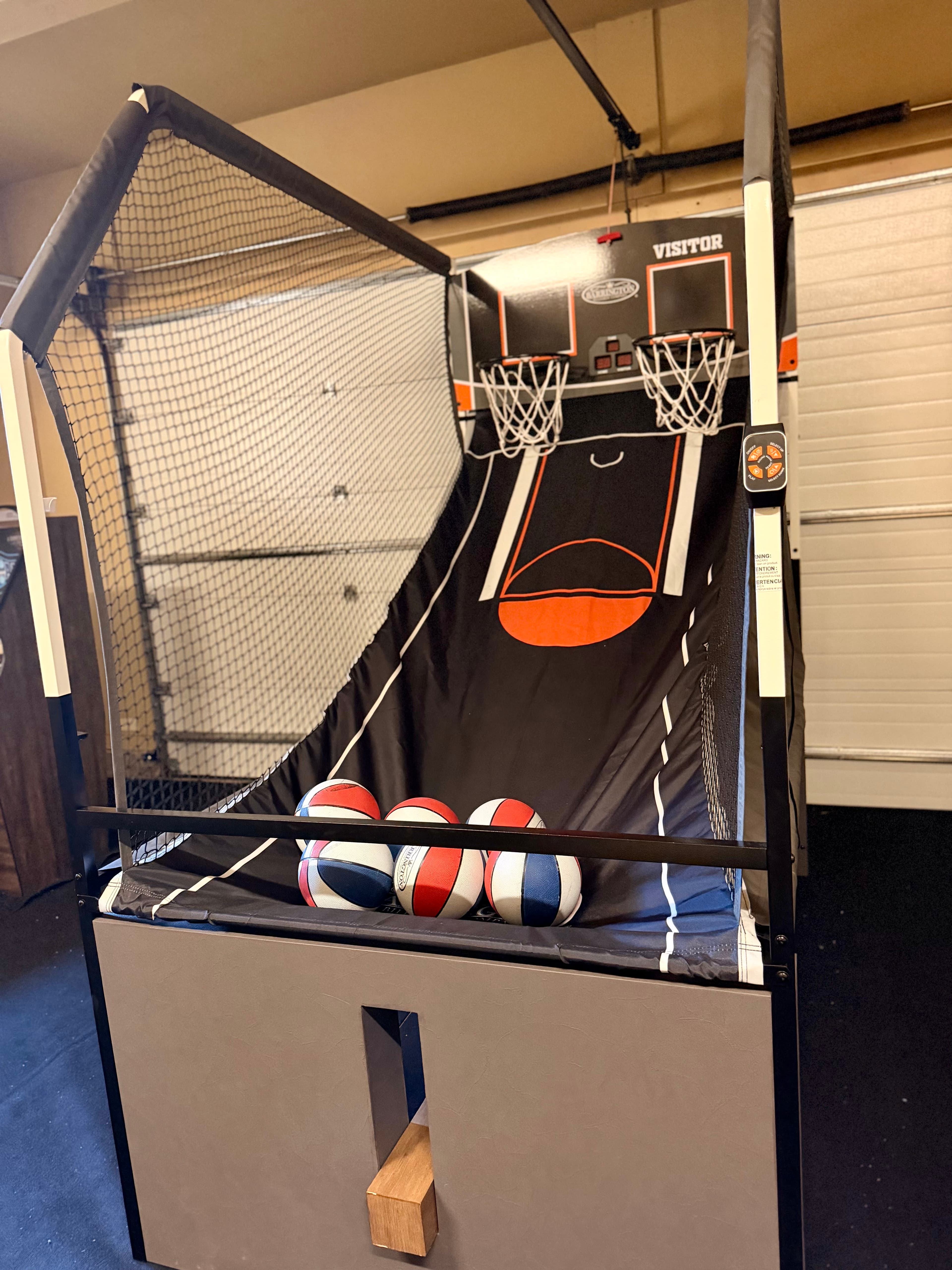 A basketball arcade game with three multi-colored balls sits against a garage wall.
