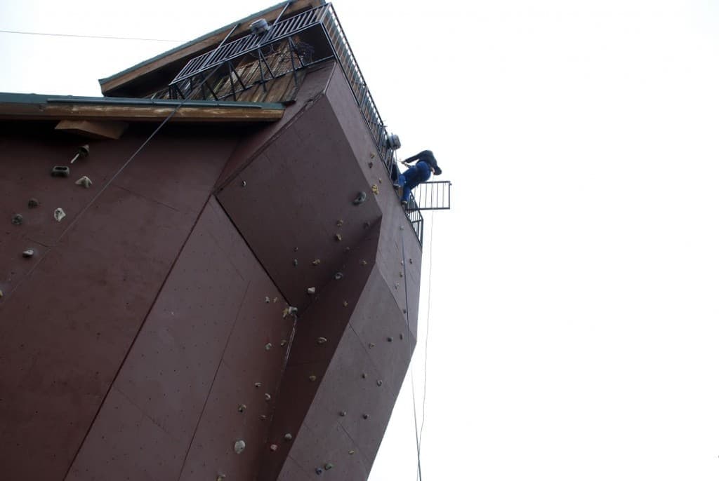 A climber ascends a large, angled rock wall with a viewing platform above.