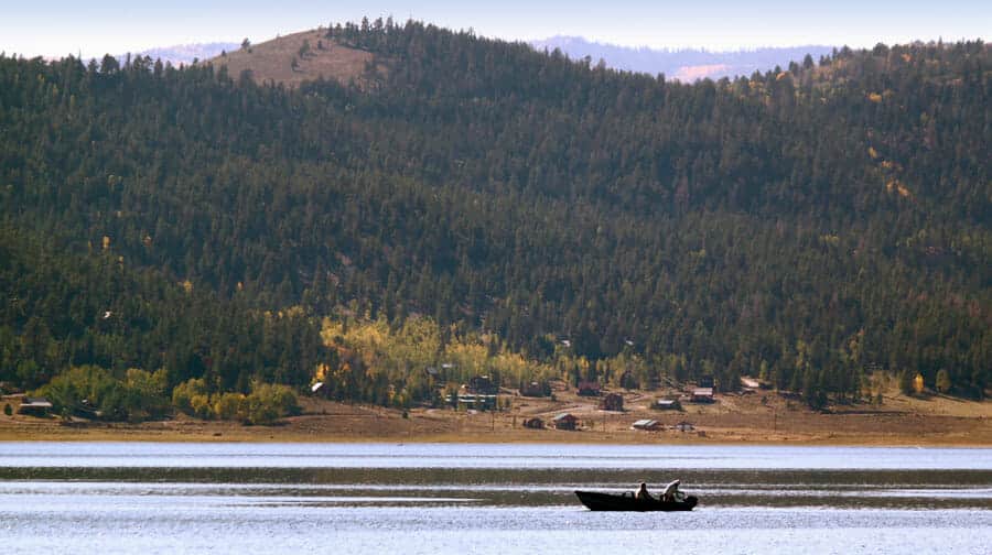 A small boat is seen on a calm lake, surrounded by forested hills and distant cabins.