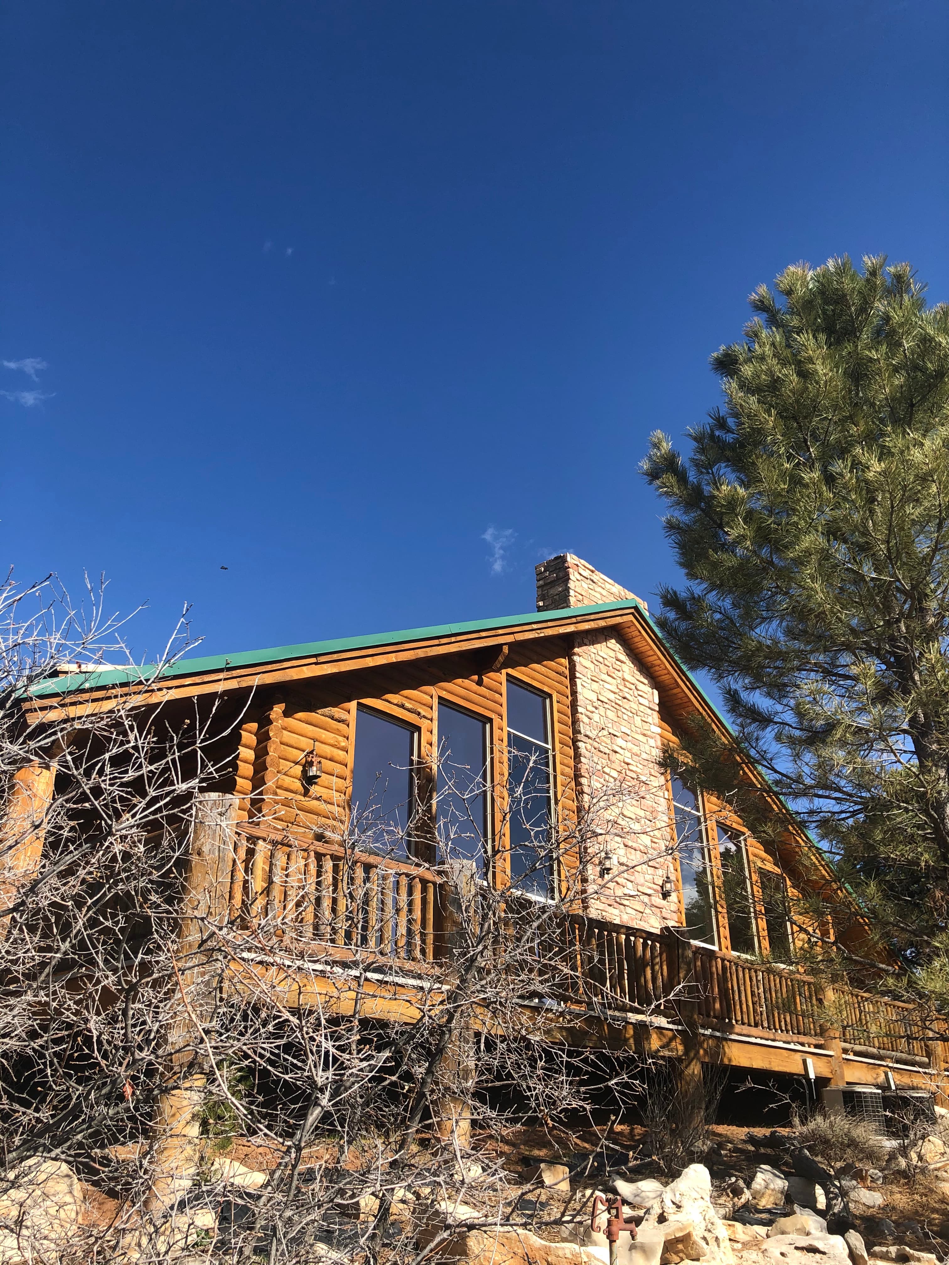 A wooden cabin with large windows and a stone chimney, surrounded by sparse vegetation under a blue sky.