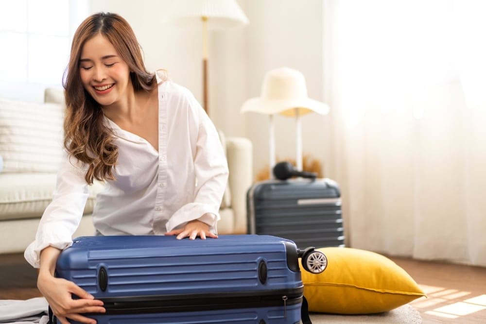 A woman smiles while packing a blue suitcase in a bright, cozy room.