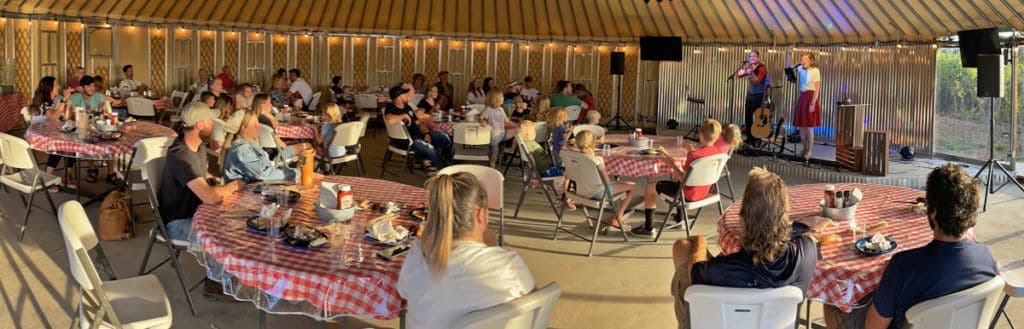 A lively gathering of people seated at tables, enjoying a meal while a performance takes place on stage.