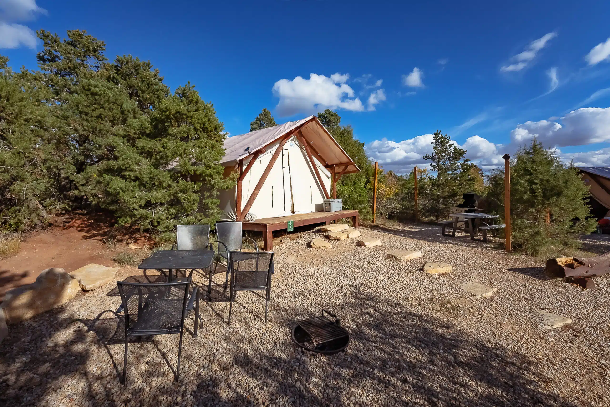 A cozy glamping tent surrounded by trees and gravel, with outdoor seating and a clear blue sky.