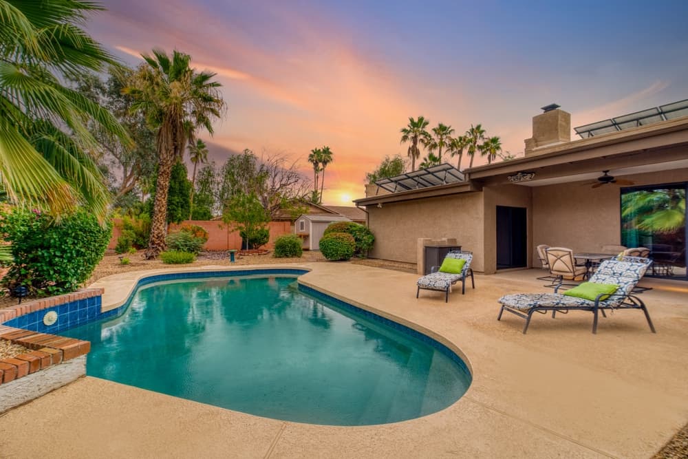 A serene backyard with a swimming pool, lounge chairs, and palm trees under a colorful sunset sky.