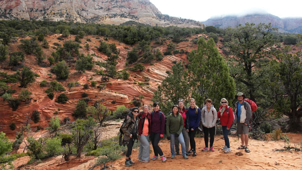 A group of eight people stands on a rocky trail against a backdrop of colorful sandstone formations and greenery.