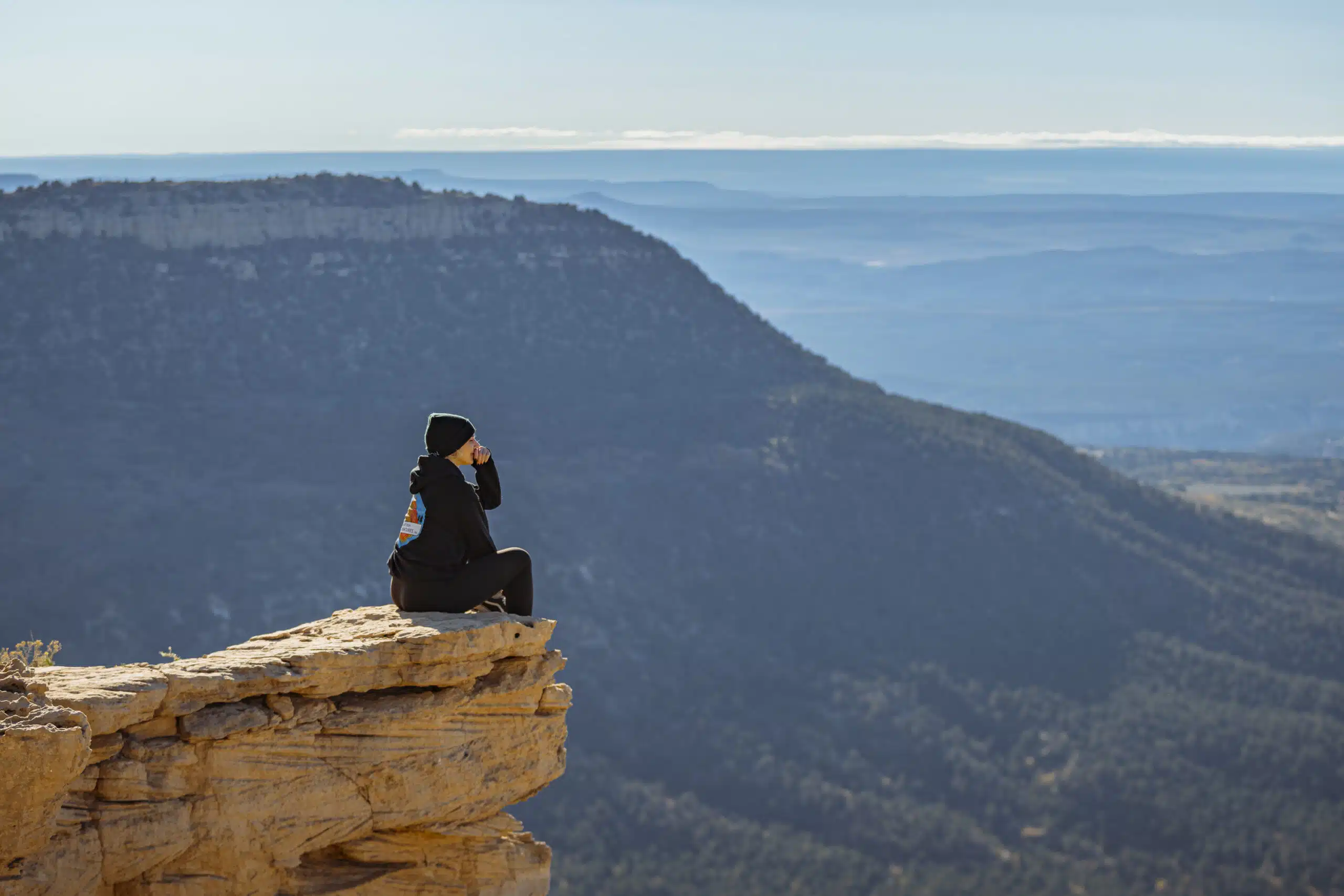 A person in a black outfit sits on a cliff, gazing at a vast landscape of mountains and valleys.